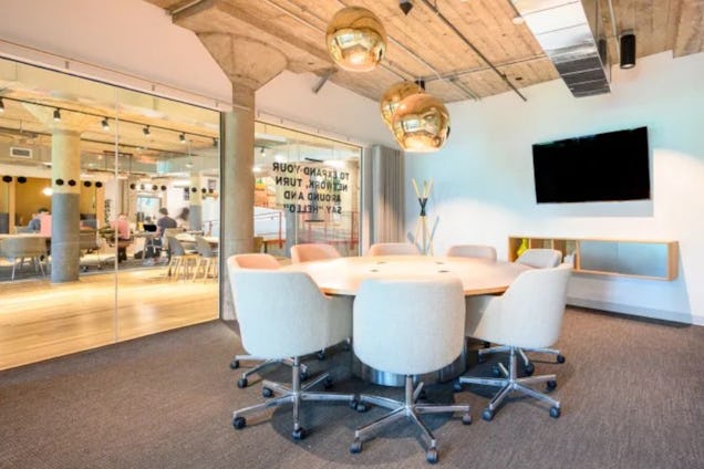 White chairs around a circular table in a meeting room with a television and glass wall.