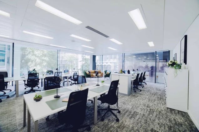 Rows of white desks with black chairs in an office with carpeted floors and glass walls.