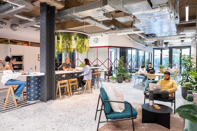 People at tables and a counter in a breakout space with chairs, plants, and exposed ceiling pipes.