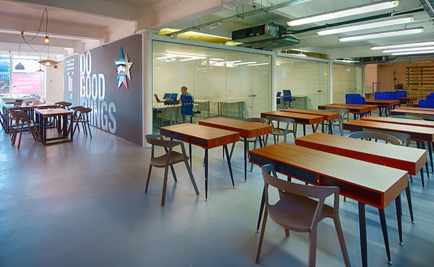 Rows of desks with chairs in a coworking space featuring a glass-walled meeting room and a logo on the wall.
