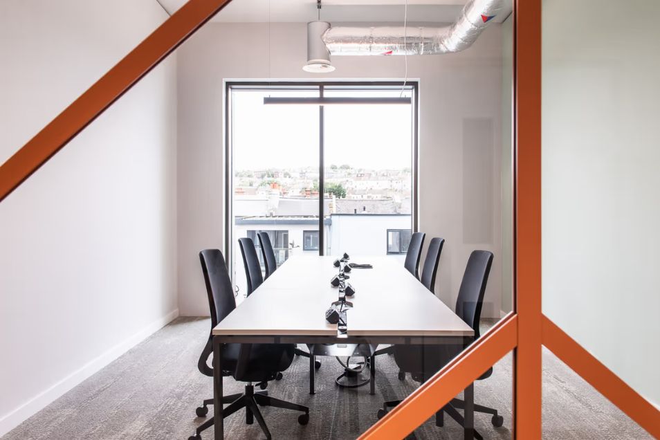 A long table with black chairs in a meeting room featuring orange beams and a window.