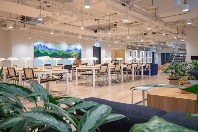 Rows of desks with chairs in a workspace featuring wood floors, plants, and a painting on the wall.