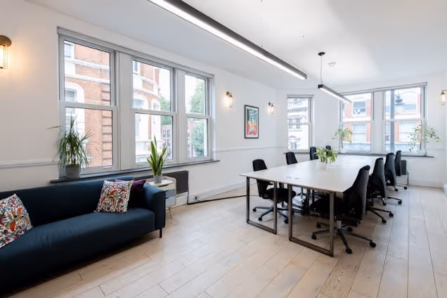 A sofa and a meeting table with chairs in a room featuring windows, plants, and wood floors.