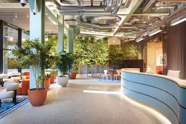 A blue reception desk in a lobby with chairs, tables, plants, and a wall of greenery.