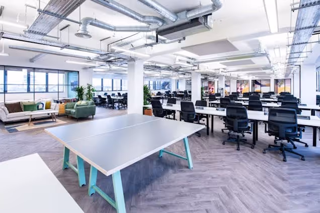 A grey and blue ping pong table in a workspace with rows of white desks, black mesh chairs, and metal pipes on the ceiling.