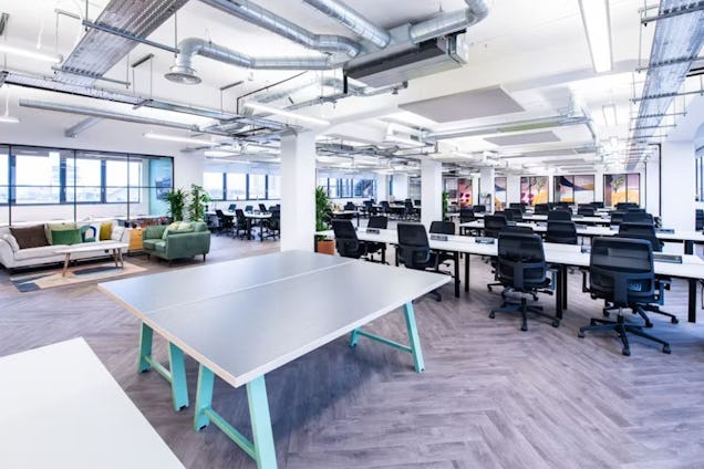 A grey and blue ping pong table in the middle of an office with rows of white desks, black mesh chairs, and ceiling pipes.
