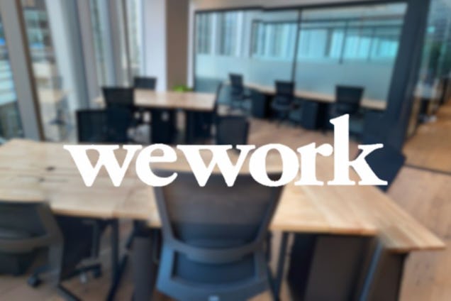 A blurry image of a wooden desk with black mesh chairs in an office setting with a "wework" logo overlaying the foreground.
