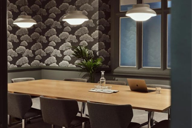 A wooden table with chairs sits under pendant lights in a meeting room with patterned wallpaper.