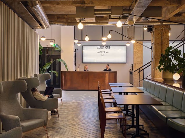 Armchairs and square metal tables with booth seating in a reception area near a wooden counter and staircase.