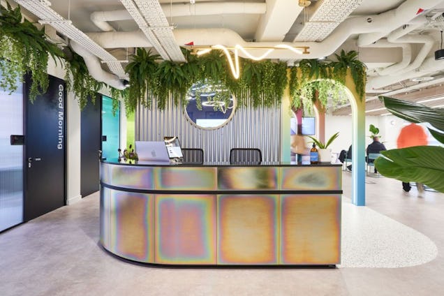 A curved reception desk stands in front of a wall with vertical slats and hanging plants.