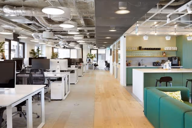 Rows of white desks with black mesh chairs sit on a wooden floor next to a green sofa in an office kitchen.