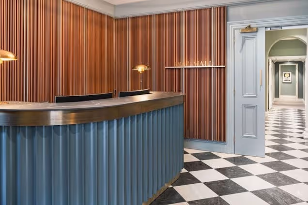 A blue reception desk stands on a checkered floor in front of wood panelling and a doorway.