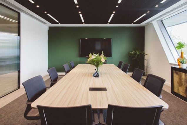Chairs surround a long wooden table in a boardroom with a wall-mounted screen and a vase of flowers.