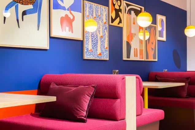 A pink sofa booths with cushions sits in front of a blue wall with framed artwork and a table.