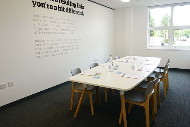 Chairs surround a white table in a meeting room with text on the wall and a window.