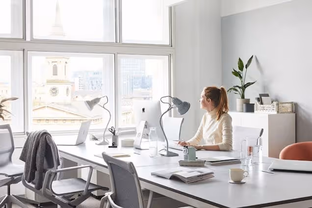 A person at a desk using a laptop in an office with windows overlooking the city.