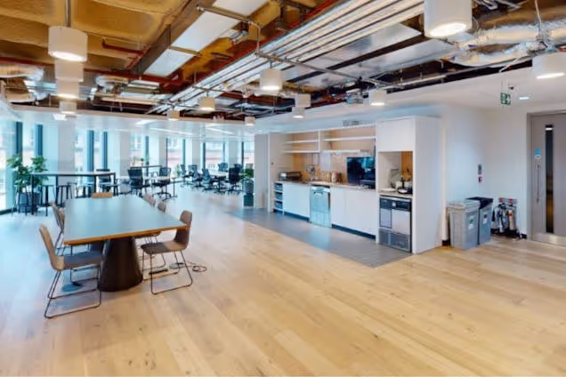 A wooden table with chairs sits on a wooden floor in a workspace with a kitchen area.