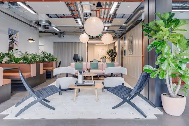 Two blue lounge chairs face a wooden table in a coworking space with a sofa, plants, globe pendant lights, and ceiling pipes.