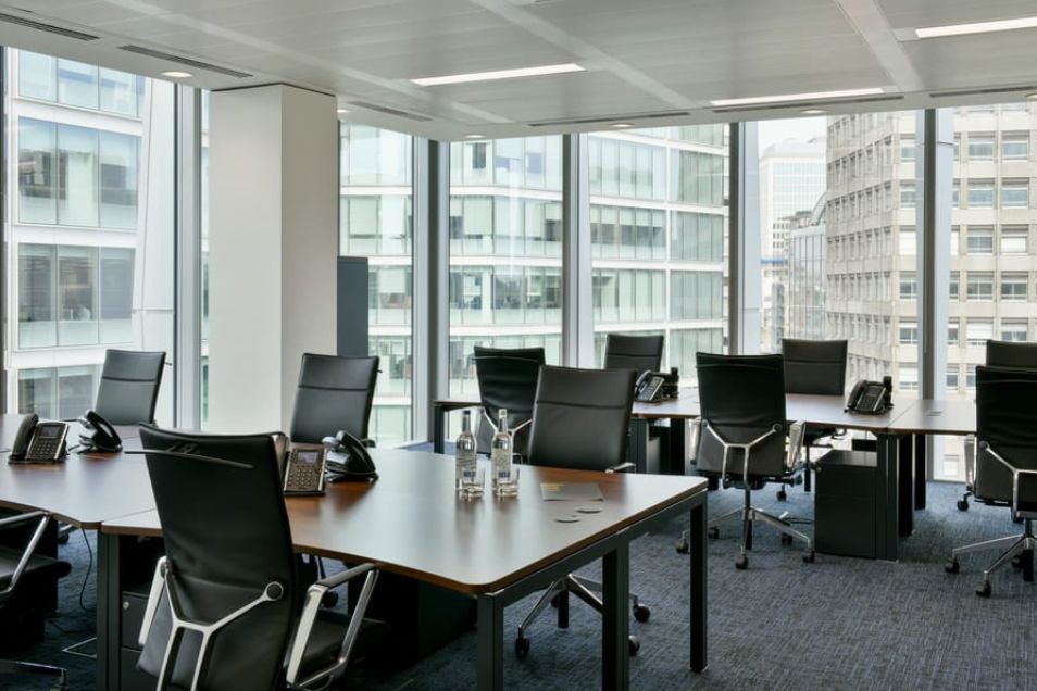 Black chairs surround a wooden table in an office with floor-to-ceiling windows and views of city buildings.