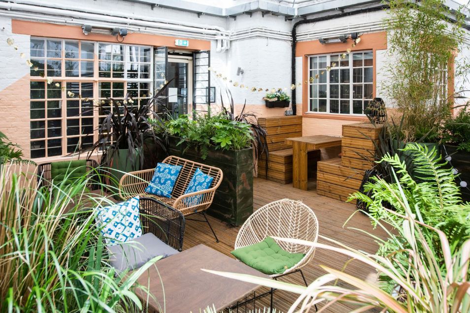 Chairs and wooden tables sit on a terrace surrounded by plants and windows.