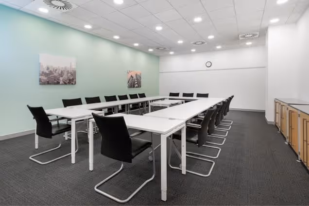 Rows of white tables and black chairs are arranged in a U-shape in a meeting room with carpet.