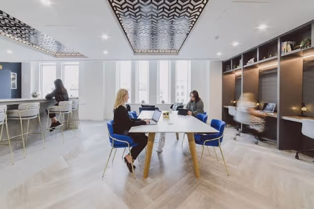 Two people sat at a white table in an office with blue chairs, a bar counter, and a herringbone floor.