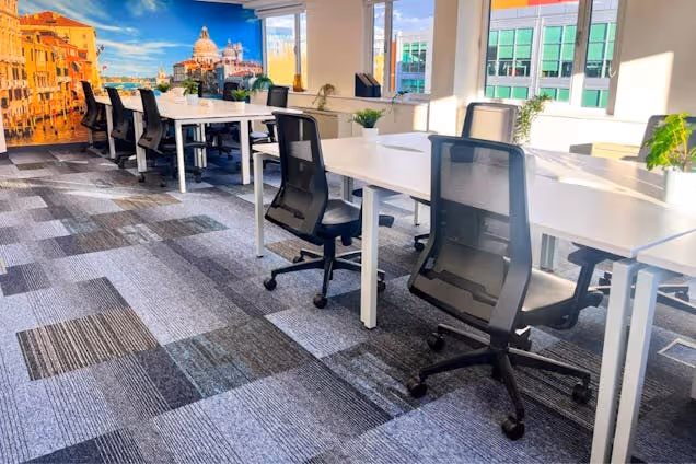 Black mesh chairs sit at white desks in an office with a mural on the wall and large windows.