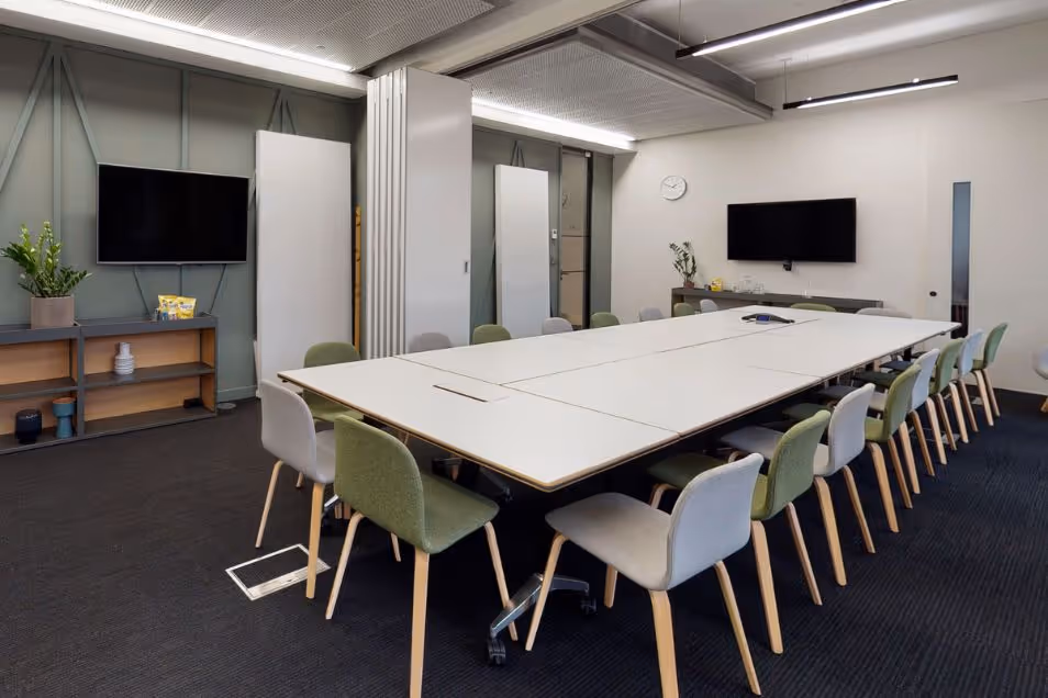 Light blue and green chairs surround a long white table in a meeting room with carpet and two screens on the wall.