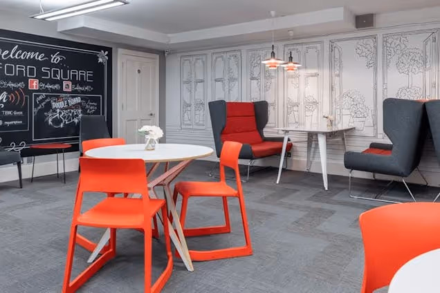 Red chairs and high-back armchairs sit around tables in a room with a chalkboard and carpet tiles.