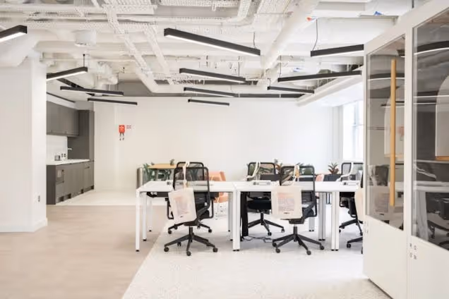 Rows of white desks with black chairs in an office with exposed ceiling pipes and white walls.