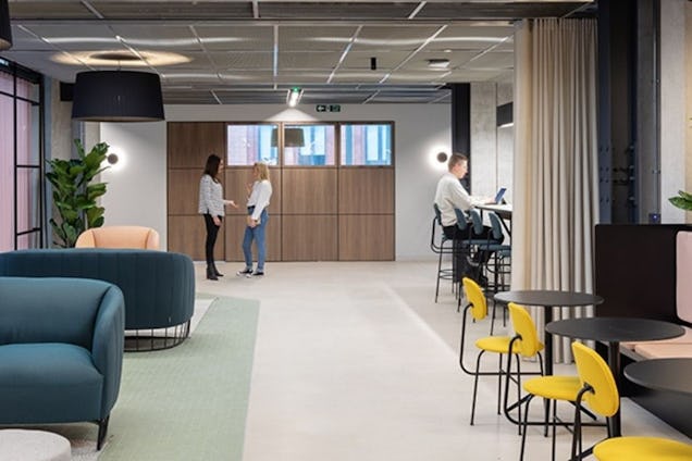Two people talking in a hallway while others sit at tables in a coworking space with teal sofas and yellow chairs.