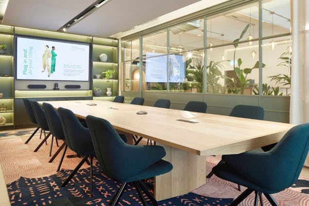 Blue chairs surround a wooden table in a boardroom with a screen and a glass wall with plants behind it.