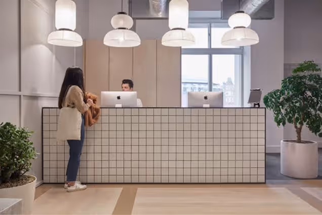 A person stands at a tiled reception desk while another person sits behind a computer monitor.