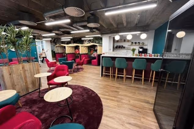 Teal stools at a kitchen island face a lounge with pink and teal chairs and light-coloured tables on a wooden floor under ceiling pipes.
