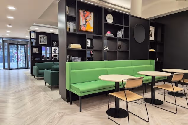 A green sofa and light wood tables with chairs in a coworking area with a black shelving unit and reception area.