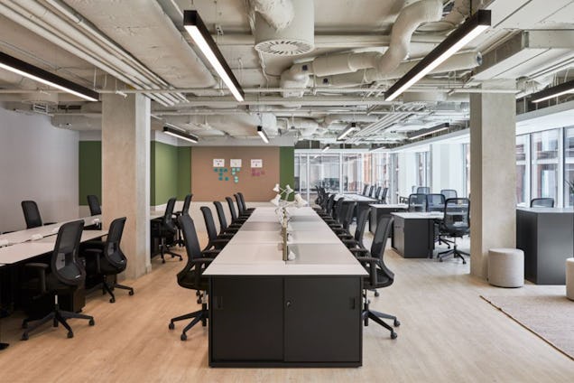 Rows of white and black desks with black chairs on a light wooden floor in an office with exposed ceiling pipes.