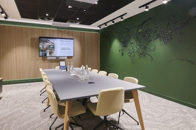 Butter yellow chairs surround a table in a meeting room with a wall-mounted screen and a wood-slat wall and a green wall.