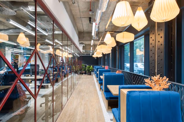 Blue booths and tables under lantern lights in a breakout space next to a glass-walled office.