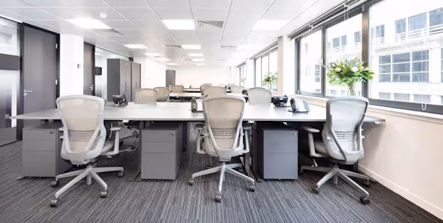 Rows of white desks with white chairs on a grey carpet floor in an office with large windows.