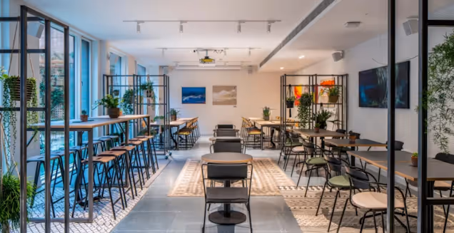 High wooden and black tables with stools and dining tables with chairs in an office cafe area with plants.