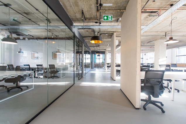 A hallway with pillars leads past glass-walled meeting rooms and open-plan desks in an office.