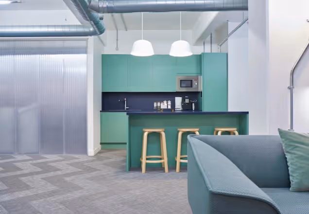 Wood stools at a green kitchen island near a sofa in an office breakout area with long dangling pendant lights.