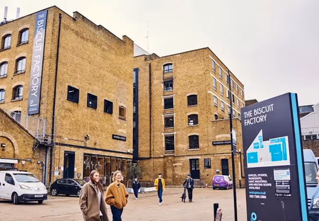 People walk on the pavement past a brick building, a direction sign, and a van under a blue sky.