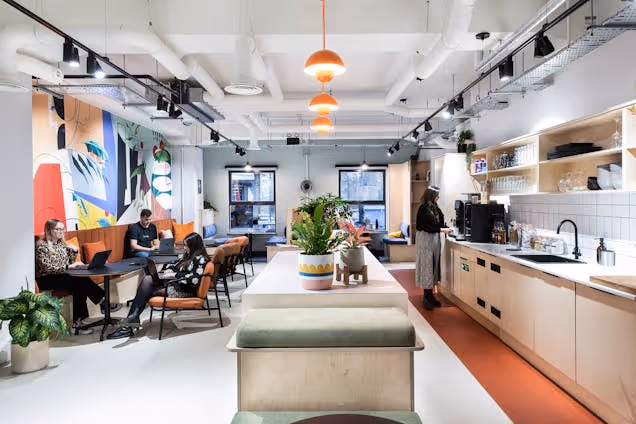 People in an office kitchen sitting on a burnt orange build in sofa in front of dark tables. There is a white kitchen island that has orange pendant lights dangling down. Another person is making coffee in the kitchen.