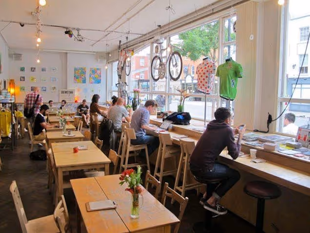 People at wooden tables and benches in a cafe with large windows and bicycles on the wall.