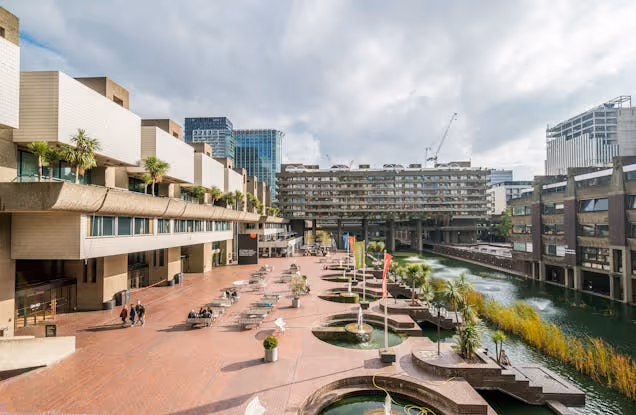 A view from above shows a courtyard with tables and people next to a pond and city buildings.