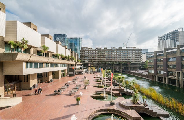 A view from above shows a courtyard with tables and people next to a pond and city buildings.