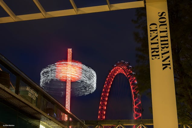 Fairground rides and a Ferris wheel glowing with red lights against a dark sky at night and a yellow metal frame.