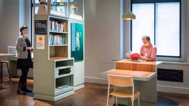 People sat at tables or looking at a bookshelf in a workspace with windows and green cabinets.