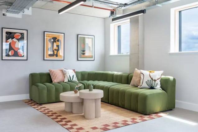 A green velvet sofa and cream abstract coffee tables on a patterned rug in a room with framed artwork and windows.